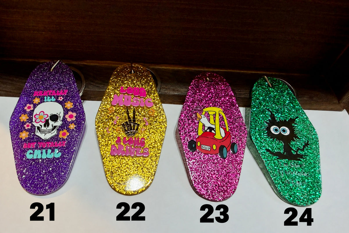 Four glittery keychains lined up on a white surface. From left to right: a purple keychain with a skull and flowers and the words 'Mentally Ill But Totally Chill'; a gold keychain with pink text reading 'Loud Music and Long Drives'; a pink keychain with a red and yellow toy car driven by a goose; and a green keychain with a black cartoon cat and the words 'I'm Fine Everything is Fine'.