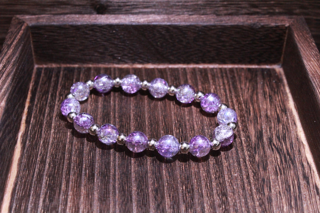 A beaded stretch bracelet made of crackle glass purple beads separated by small silver spacer beads, displayed inside a dark brown wooden tray.