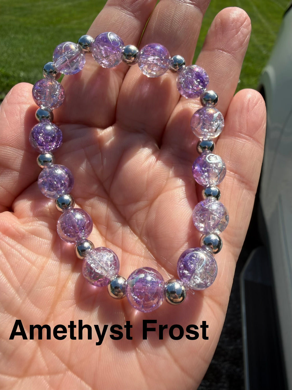 A beaded stretch bracelet made of translucent lavender-purple crackle glass with small silver spacer beads, shown resting on an open hand outdoors. The text 'Amethyst Frost' is overlaid at the bottom.
