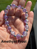 A beaded stretch bracelet made of translucent lavender-purple crackle glass with small silver spacer beads, shown resting on an open hand outdoors. The text 'Amethyst Frost' is overlaid at the bottom.