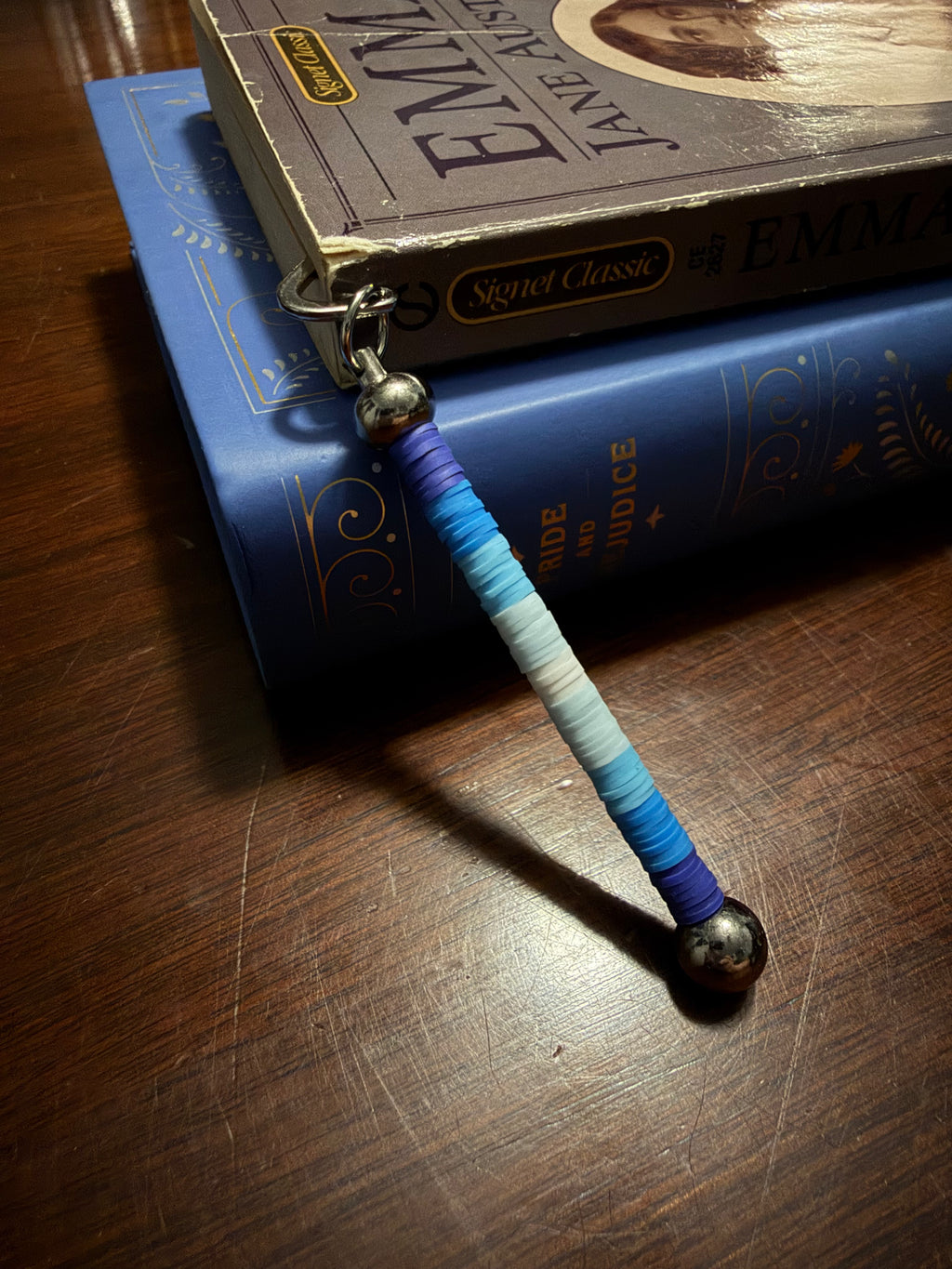 A beaded bookmark with a gradient of blue and white heishi clay beads, finished with silver metal beads and a silver split ring, resting on a stack of books on a wooden table.