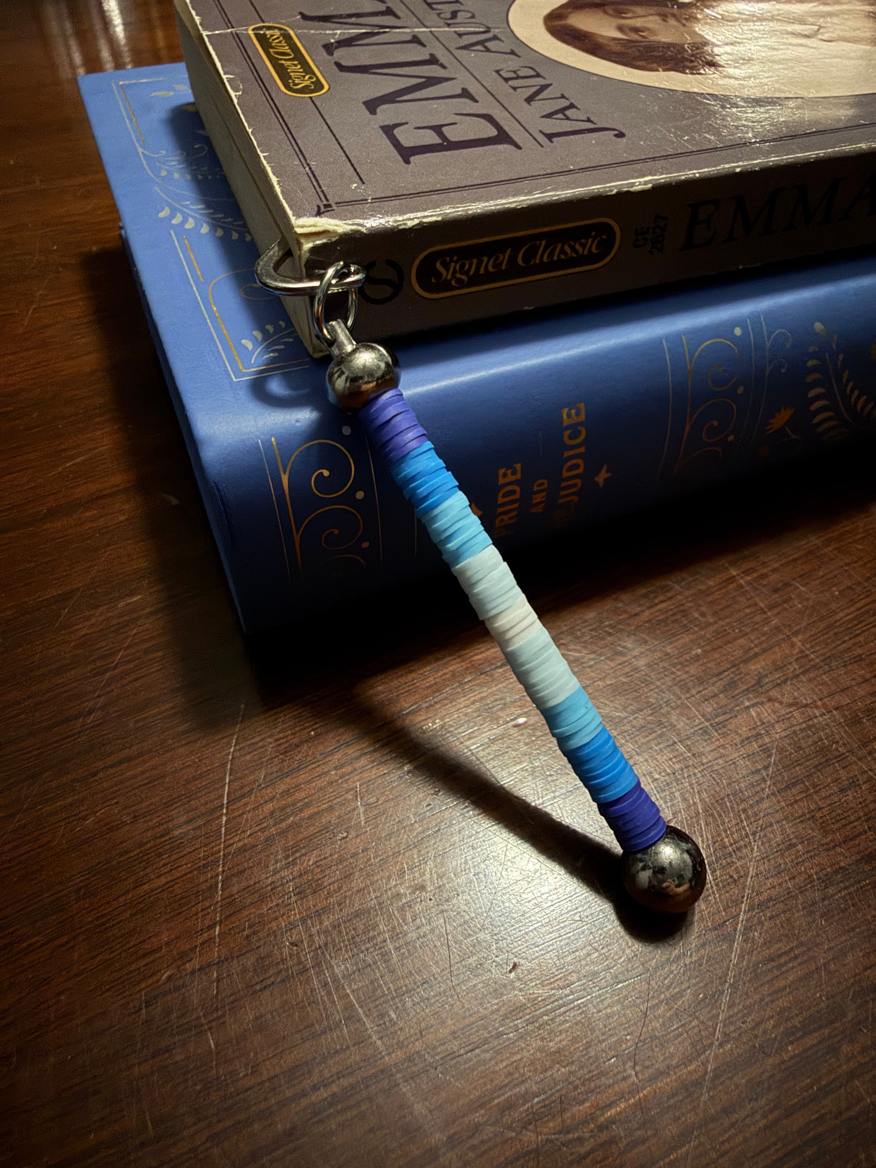 A beaded bookmark with a gradient of blue and white heishi clay beads, finished with silver metal beads and a silver split ring, resting on a stack of books on a wooden table.