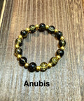 A beaded stretch bracelet featuring black and gold crackle glass beads, displayed on a brown wooden surface with the text 'Anubis' printed below.