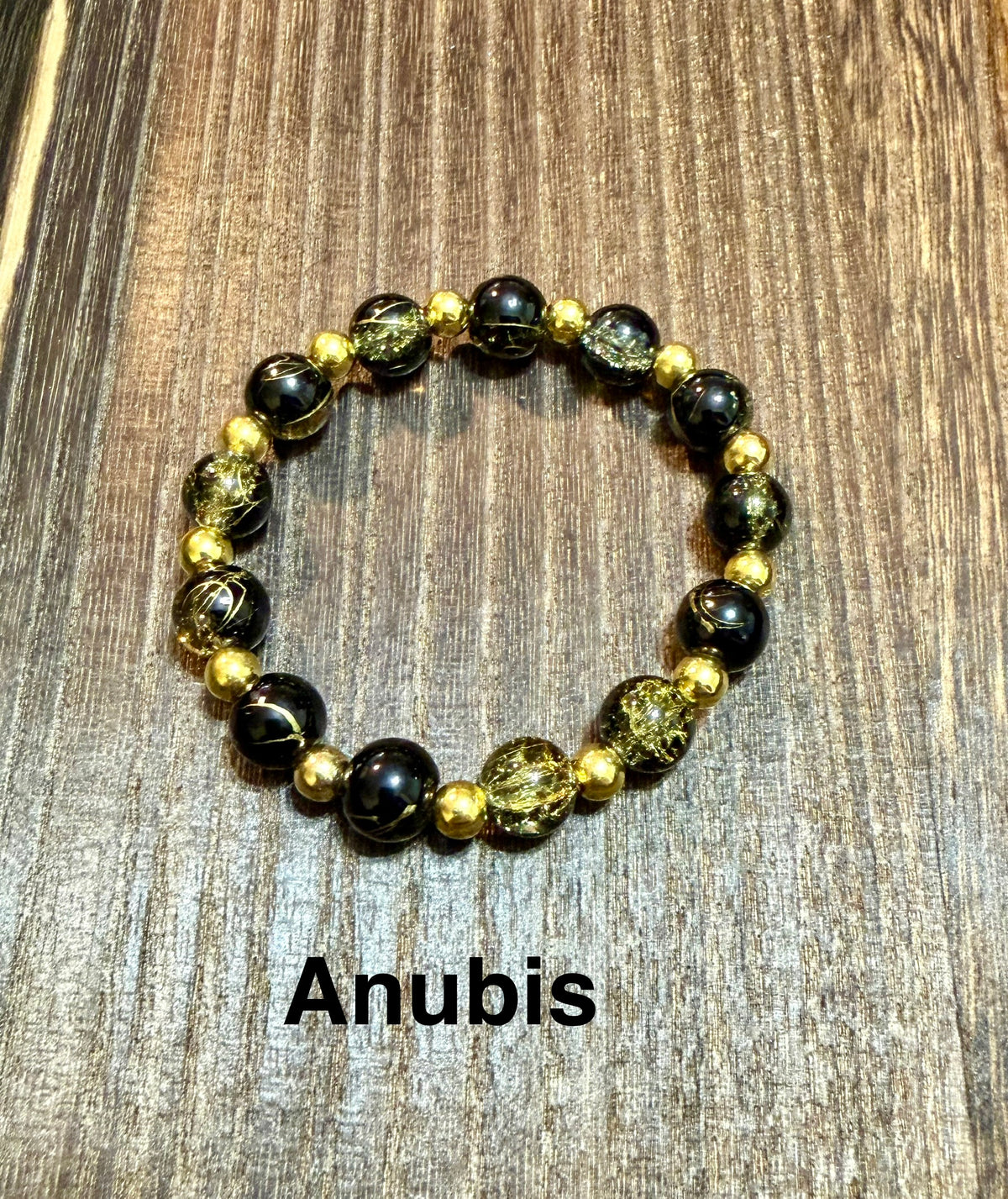 A beaded stretch bracelet featuring black and gold crackle glass beads, displayed on a brown wooden surface with the text 'Anubis' printed below.