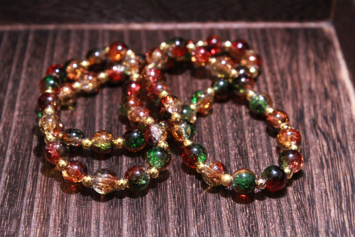 Three beaded stretch bracelets with crackle glass beads in green, red, amber, and gold, arranged on a dark wood surface.