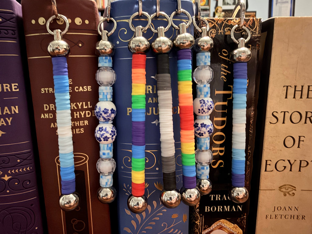 A set of colorful beaded keychains with silver metal rings and ball charms, hanging in front of a row of decorative books.