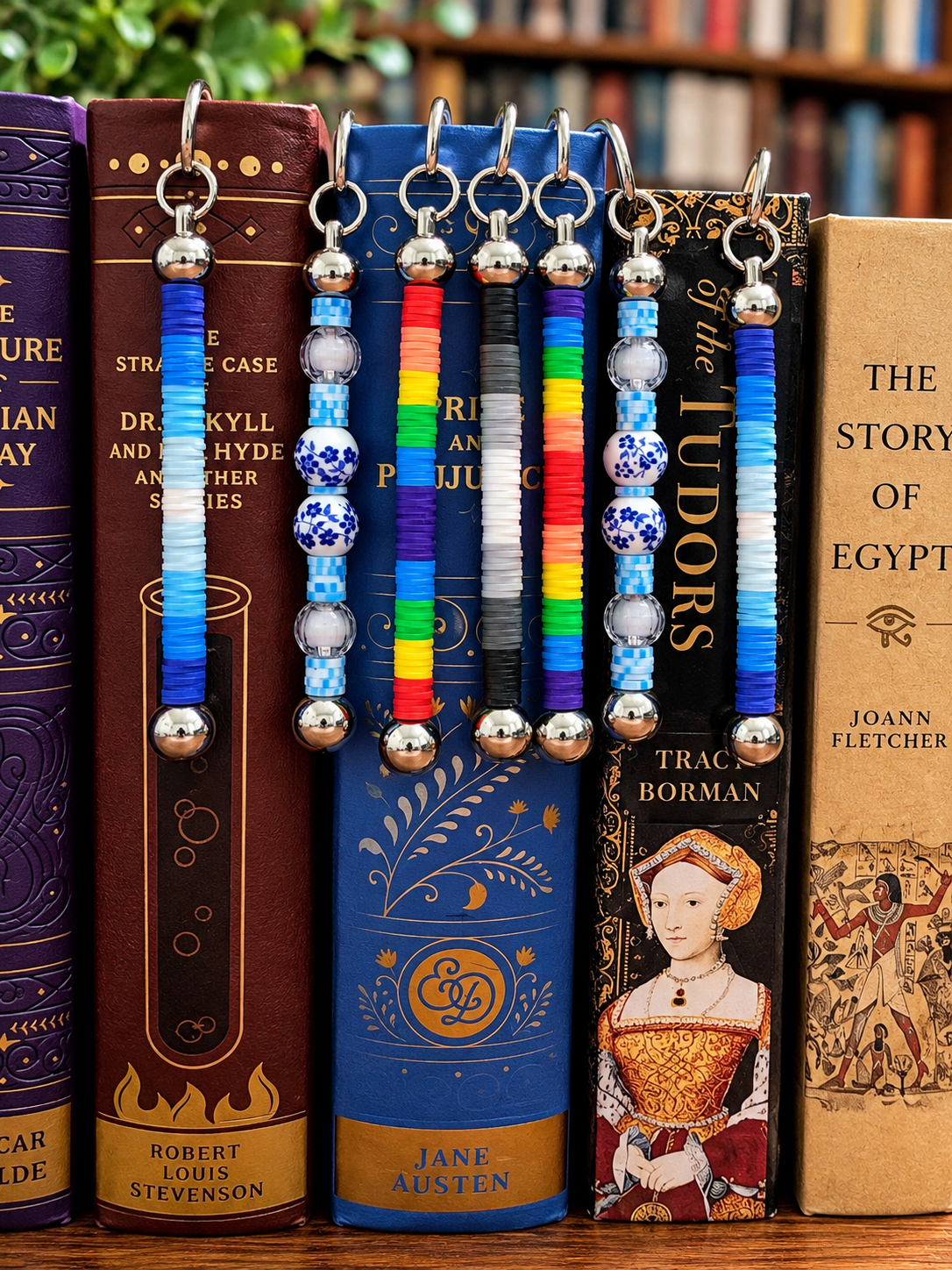 Seven beaded book bookmarks in stainless steel are shown hanging from the spines of various classic book spines, including Robert Louis Stevenson's Dr. Jekyll and Hyde and Jane Austen's Pride and Prejudice.