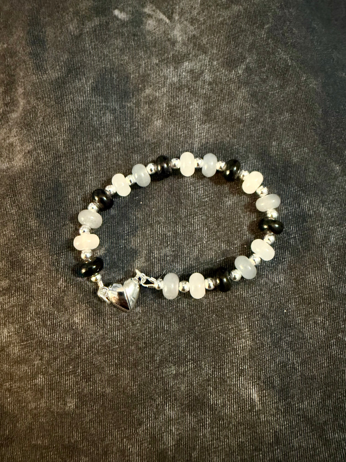 A beaded bracelet featuring black, grey, and white beads, silver spacer beads, and a small silver heart clasp, displayed on a dark textured fabric background.