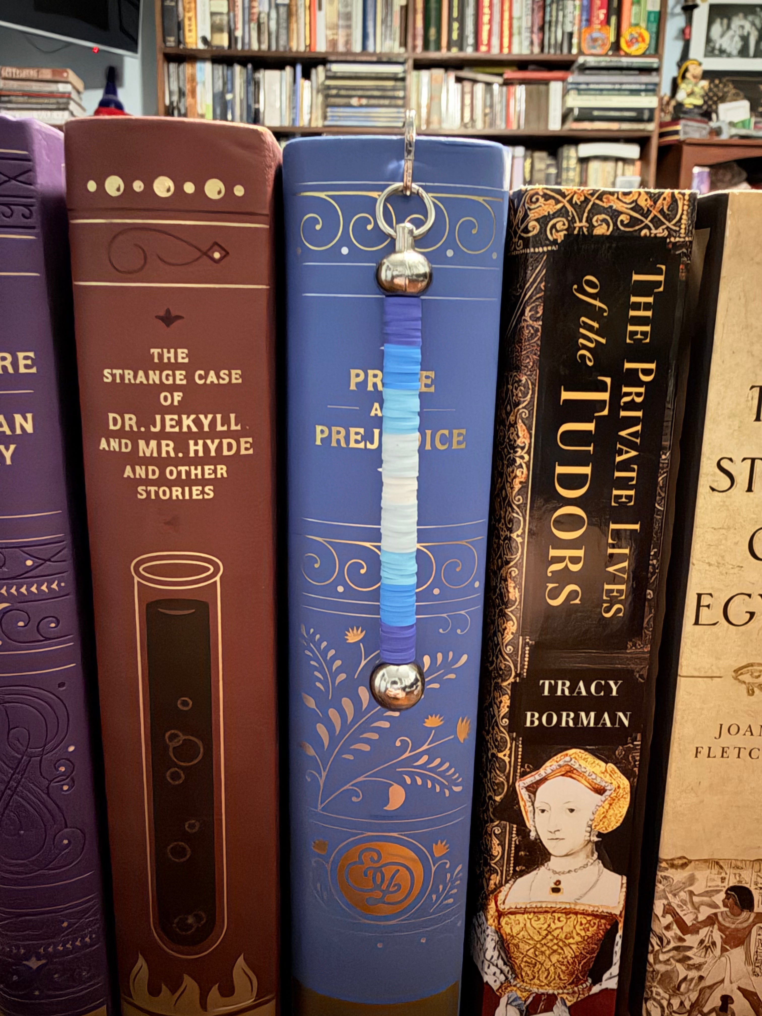 A close-up of a decorative blue beaded bookmark against book spines in a library or bookstore setting, including titles like 'The Strange Case of Dr. Jekyll and Mr. Hyde.'
