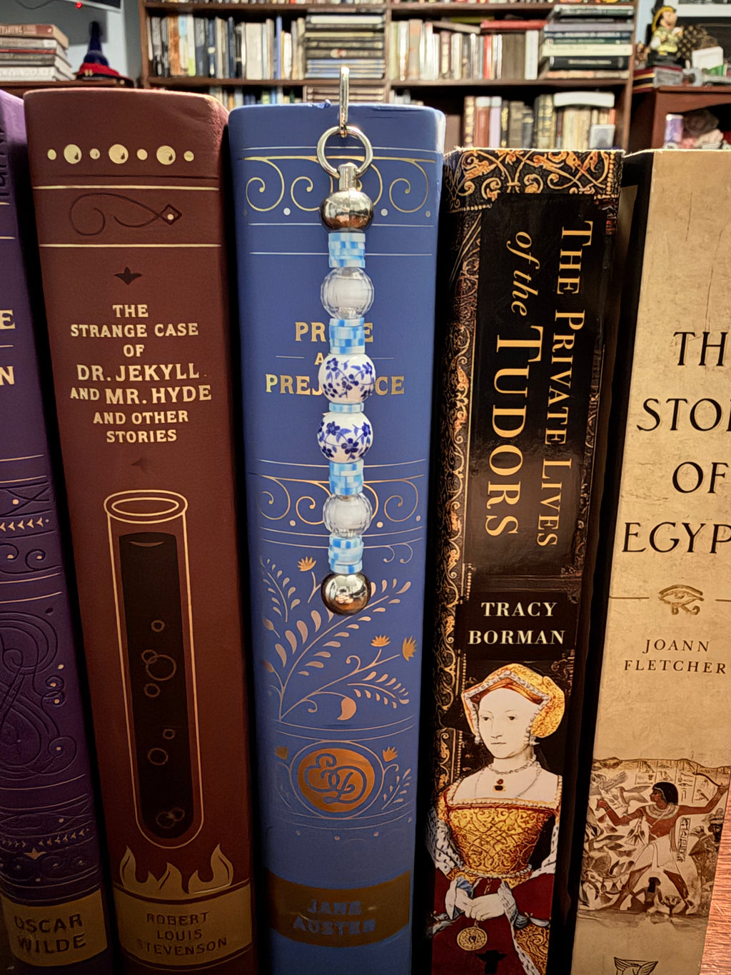 A decorative book bookmark with blue and white patterned beads hangs from the spine of a blue book on a bookshelf, surrounded by other books.