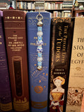 A decorative book bookmark with blue and white patterned beads hangs from the spine of a blue book on a bookshelf, surrounded by other books.