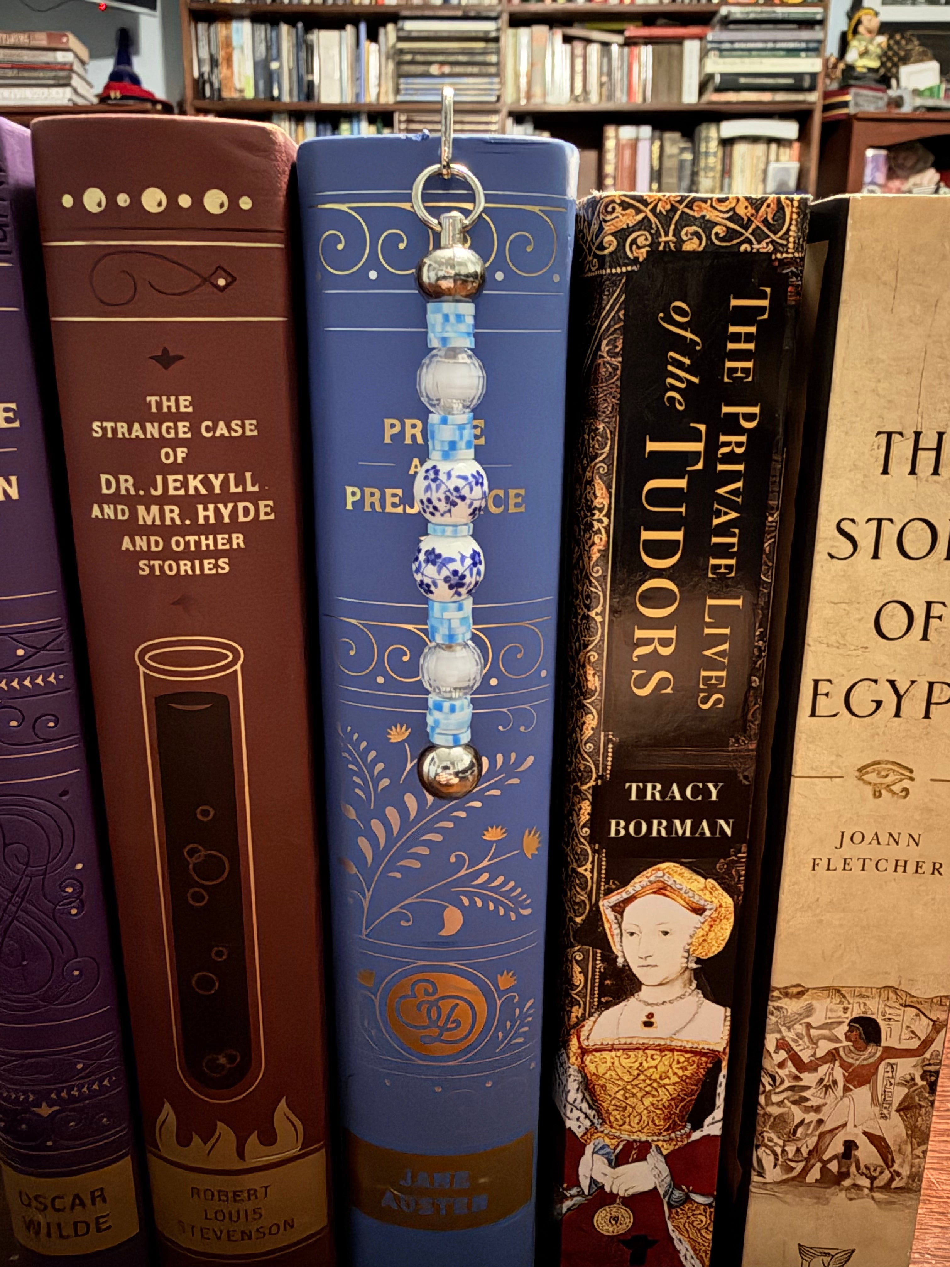 A decorative book bookmark with blue and white patterned beads hangs from the spine of a blue book on a bookshelf, surrounded by other books.