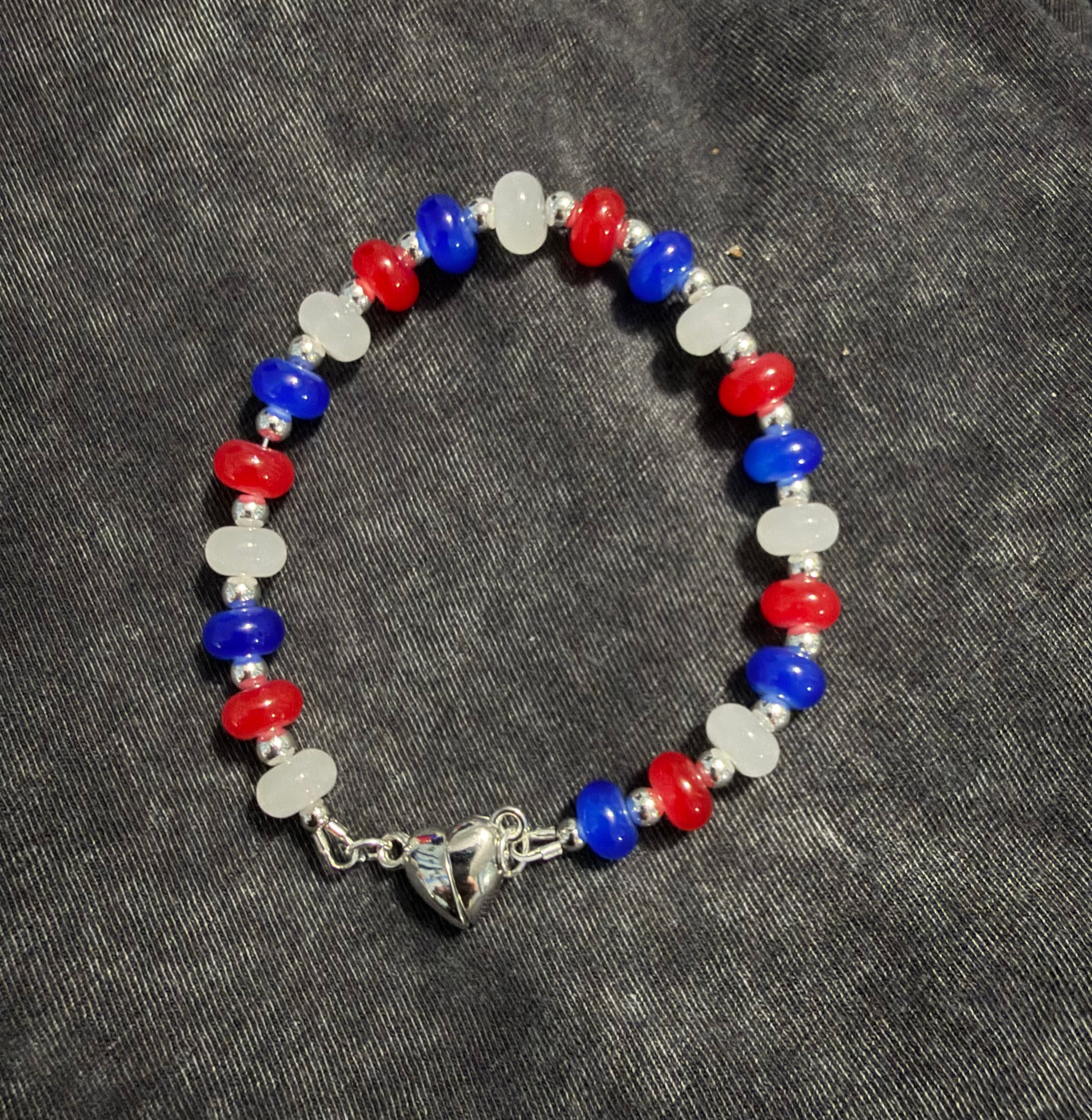 A beaded stretch bracelet featuring alternating red, blue, and white beads, finished with a silver-toned heart clasp, displayed on a dark gray textured fabric background.