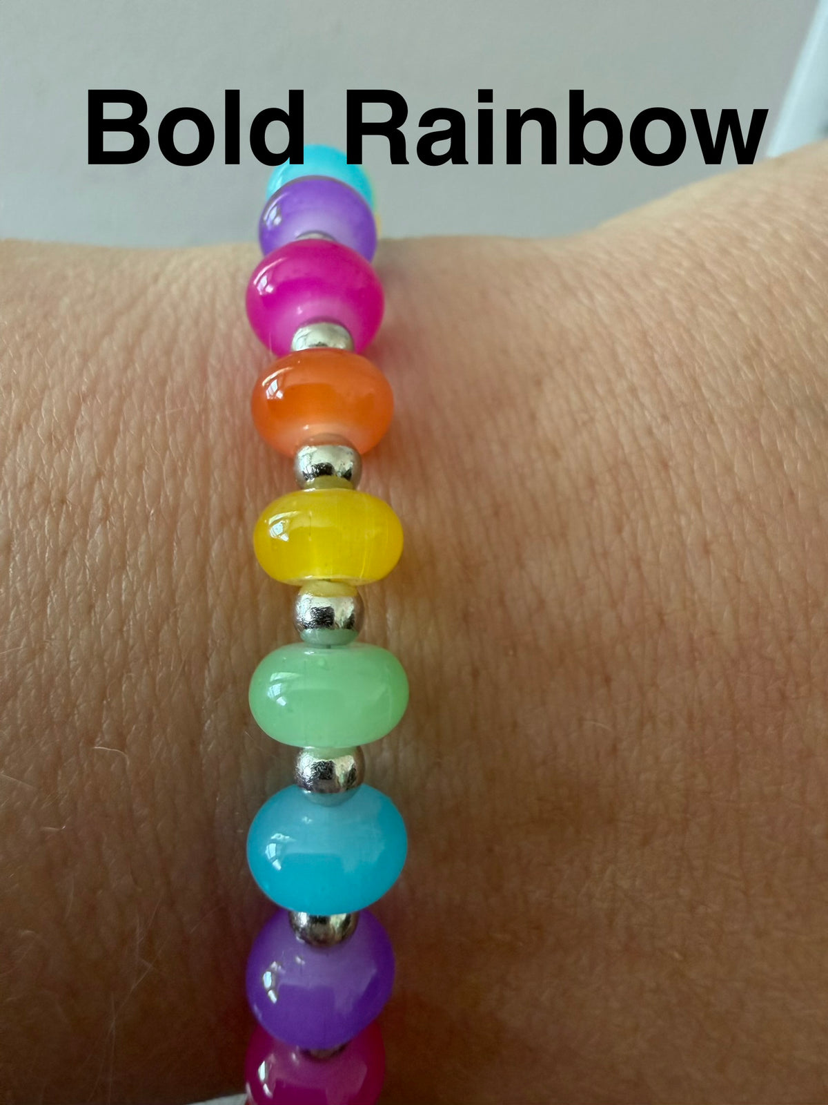 A close-up of a beaded stretch bracelet worn on a wrist, featuring vibrant rainbow-colored translucent beads separated by small silver spacer beads. The text 'Bold Rainbow' is overlaid at the top.