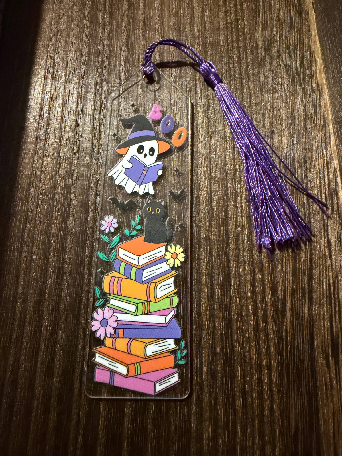 A Halloween acrylic bookmark with a reading ghost, bats, a black cat, and flowers on a stack of colorful books.  It has a matching purple tassel.