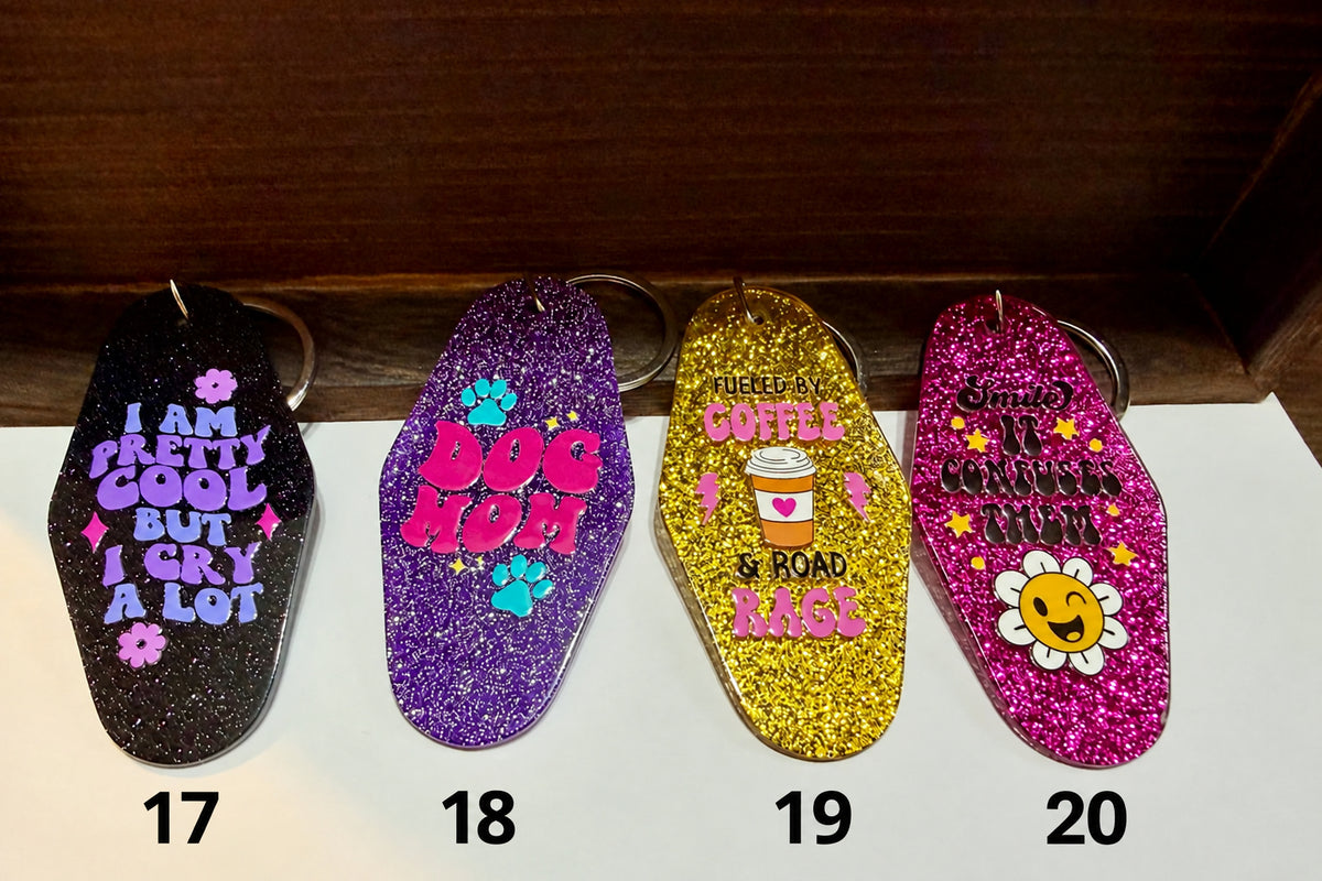 Four coffin-shaped glitter keychains lined up on a white surface against a dark wood background. Each keychain has a different colorful graphic and text: one black with purple text, one purple with pink text and paw prints, one gold with yellow text, and one pink with a smiling flower graphic.