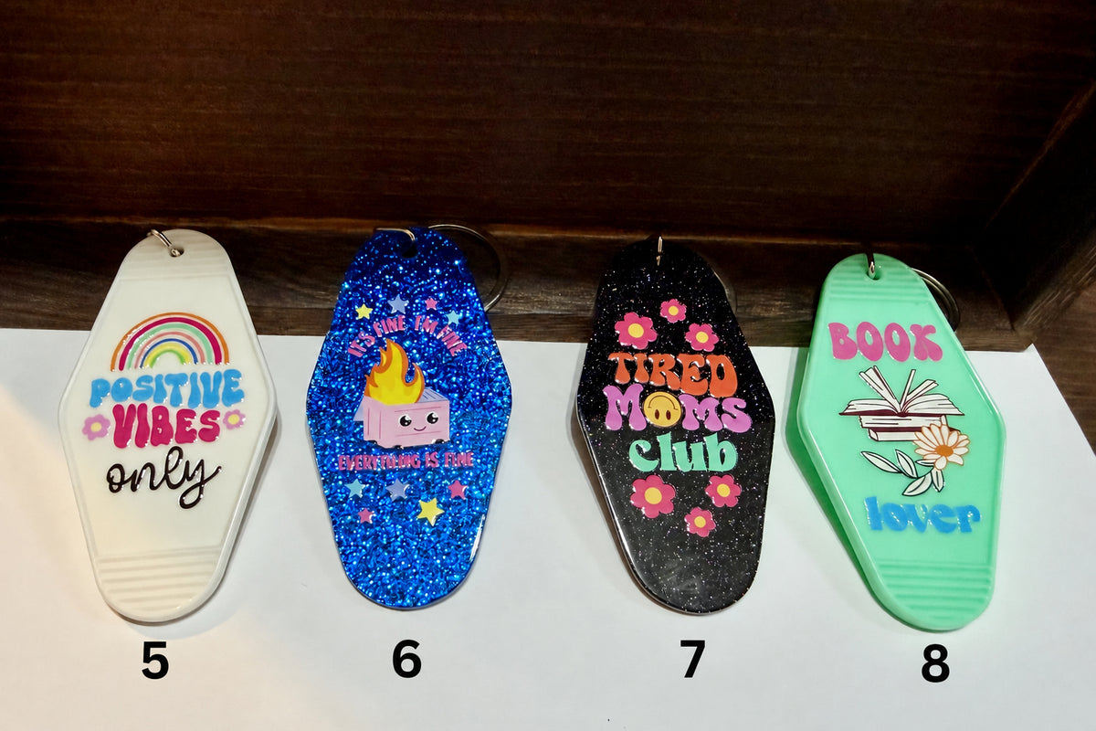Four coffin-shaped keychains lined up on a white surface against a dark wood background. The keychains feature different colorful designs: white with a rainbow and 'Positive Vibes Only', blue glitter with a cartoon character and 'It's Fine I'm Fine Everything is fine', black glitter with 'Tired Moms Club' and flowers, and mint green with 'Book Lover' and an open book illustration.