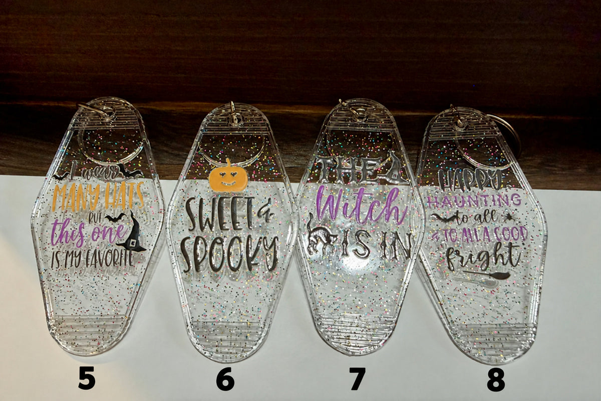 Four clear plastic Halloween-themed keychains with glitter and printed text, displayed on a white surface with a dark wooden background.  Sayings are "I wear many hats but this one is my favorite" "sweet and spooky" "the witch is in" and "happy haunting to all and to all a good fright"