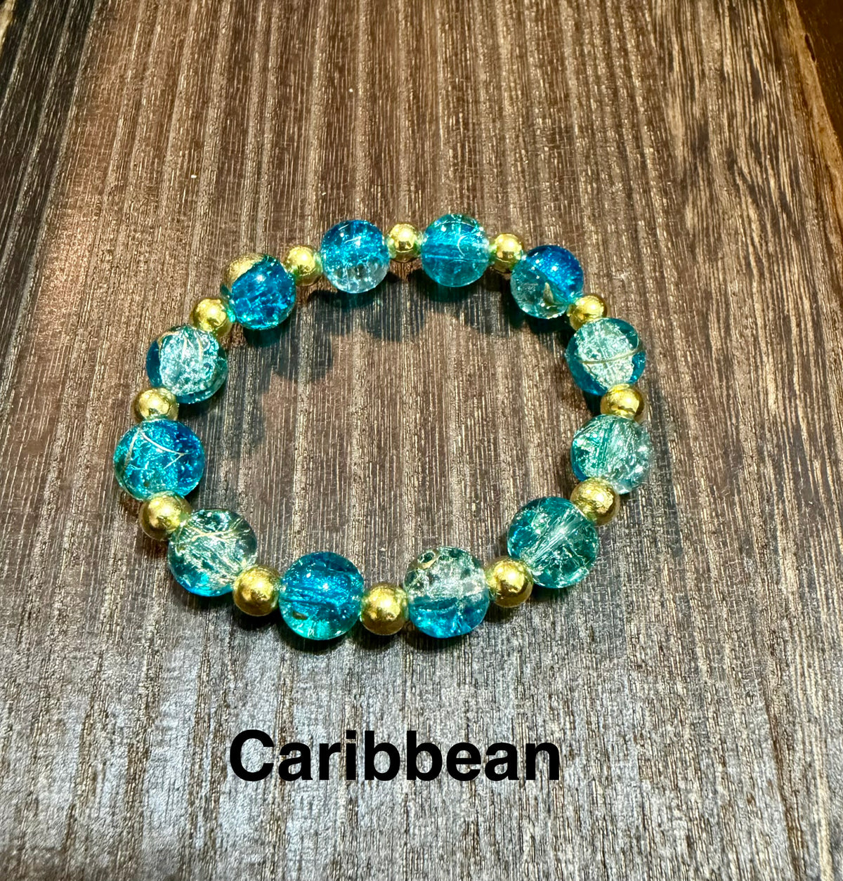 A beaded stretch bracelet with turquoise-blue crackle glass beads and small metallic gold spacer beads, displayed on a brown wood surface with the text 'Caribbean' underneath.