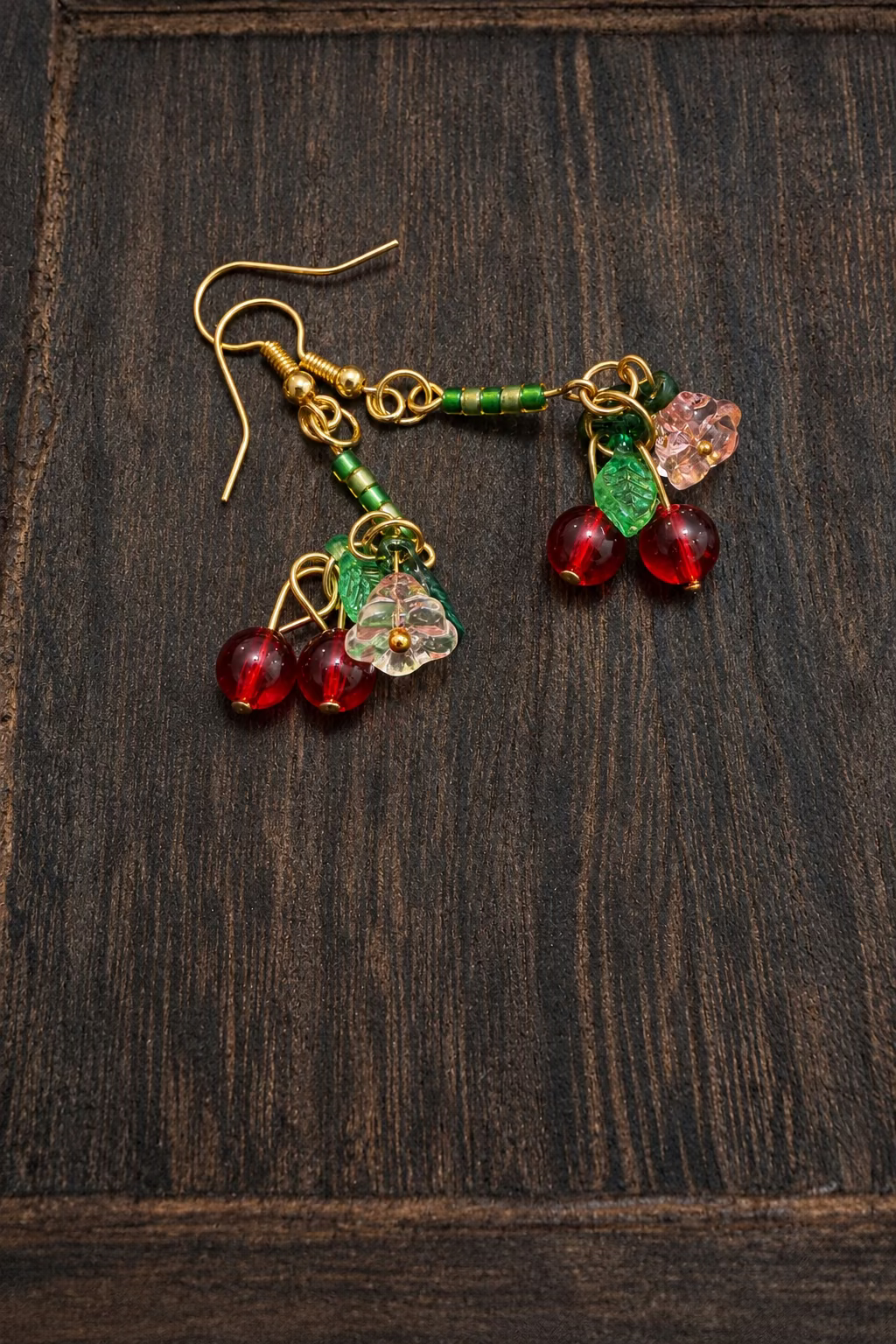 A pair of gold-tone dangle earrings with green beaded stems, light pink flower charms, and red bead spheres, displayed on a dark wood surface.