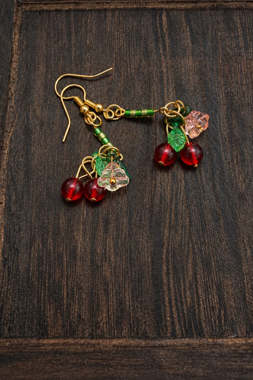 A pair of gold-tone dangle earrings with green beaded stems, light pink flower charms, and red bead spheres, displayed on a dark wood surface.