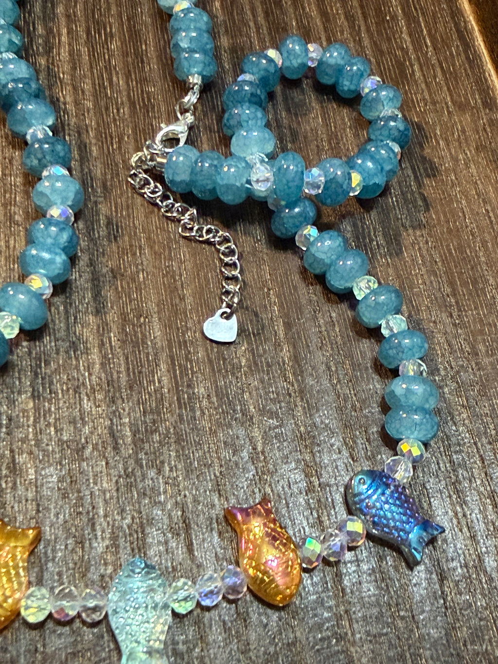 A beaded necklace with light blue aquamarine stone beads, clear iridescent faceted beads, and fish-shaped glass beads in blue, orange, and green, displayed on a dark wood surface with a heart extender chain dangling downward.