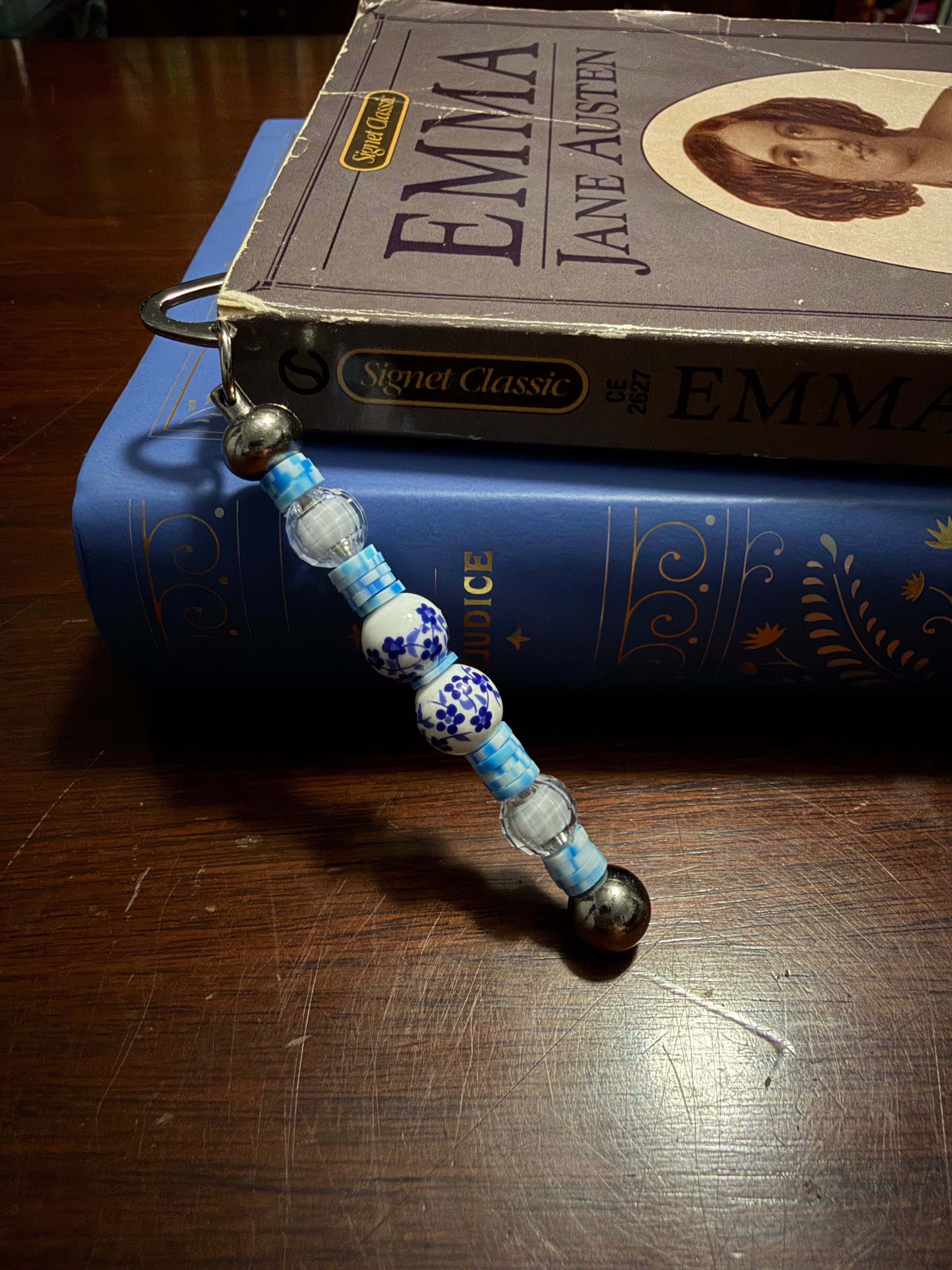 A beaded book charm with blue and white patterned beads and silver-toned metal rings, resting on top of a blue Signet Classic book which is stacked on another book on a wooden table.