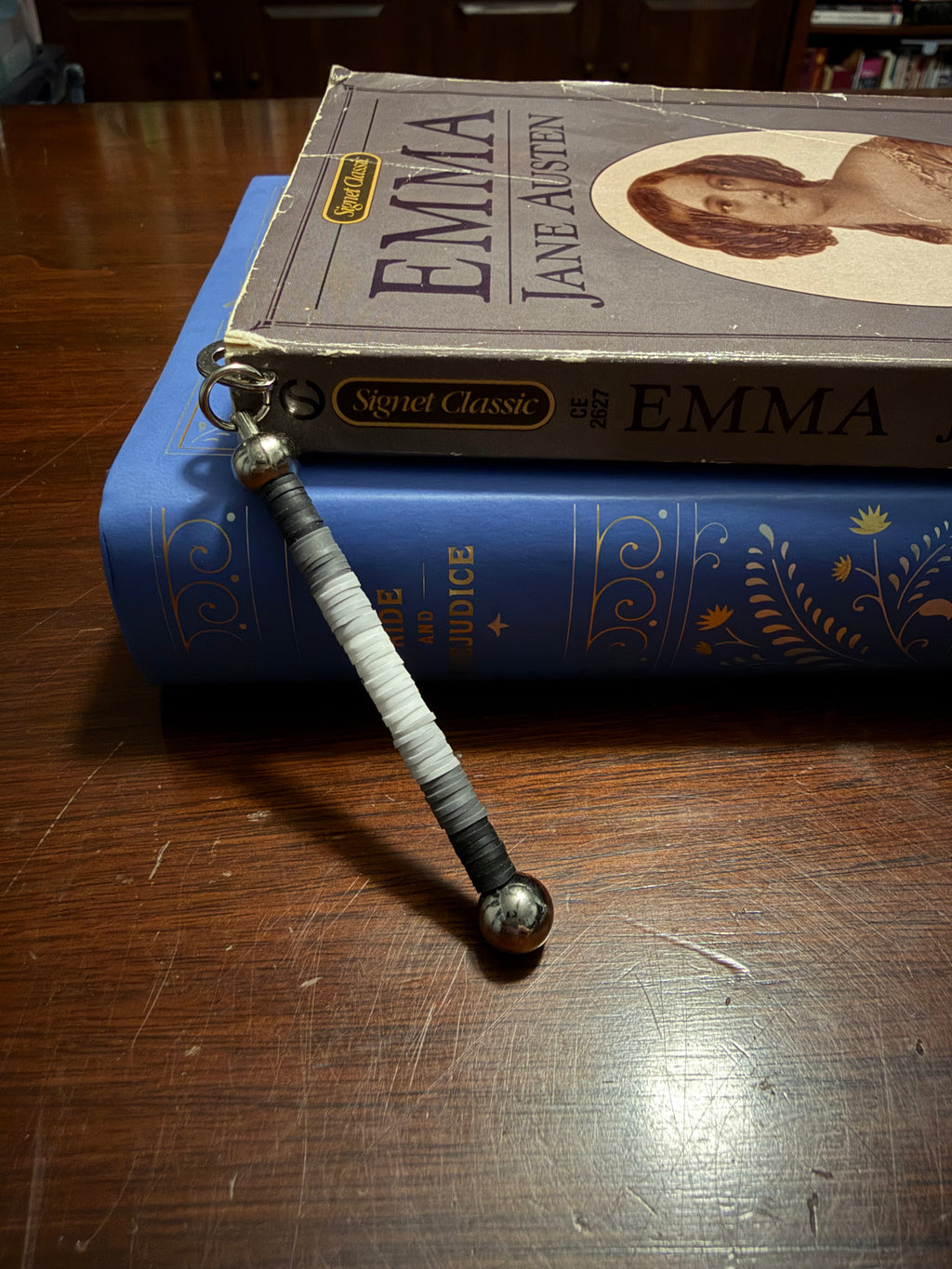 A beaded book charm with a striped black, white, and gray cord hangs from a metal ring, resting on top of a blue book and a vintage copy of Emma on a wooden table.