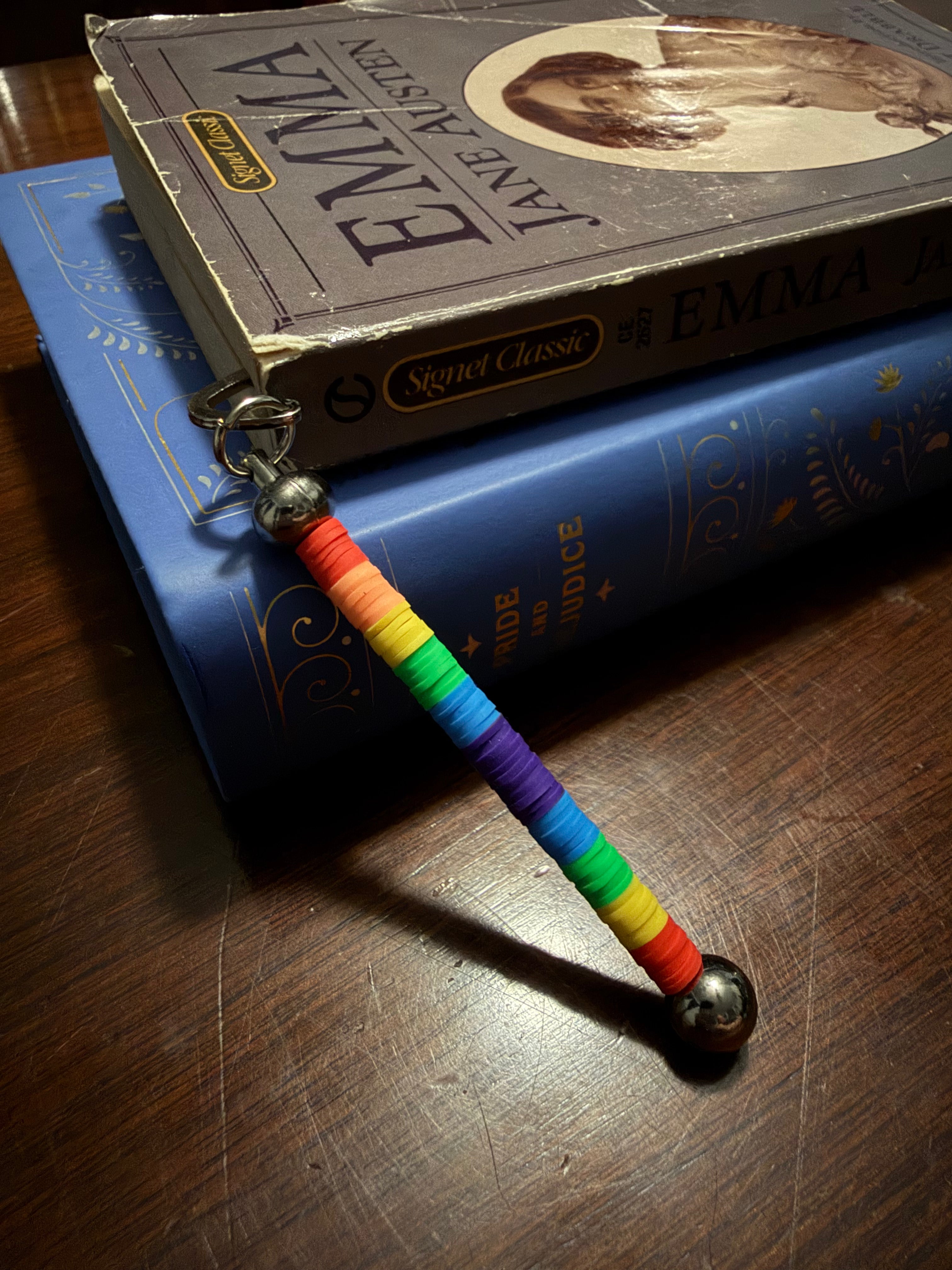 A rainbow-striped beaded bookmark with silver metal rings and balls, resting on a blue book which is stacked on top of a vintage Jane Austen book on a wooden table.