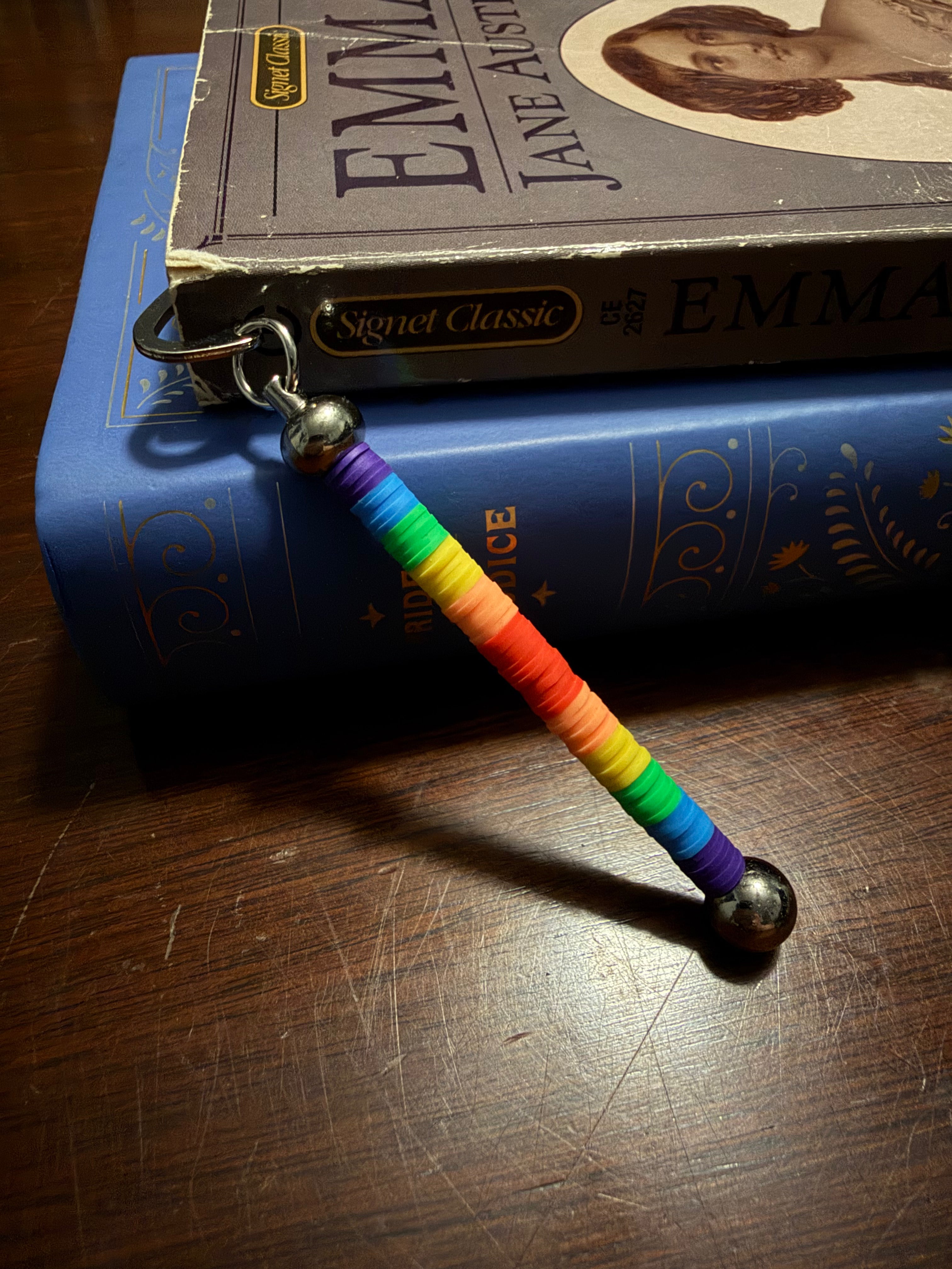 A rainbow beaded bookend with silver metal balls at the ends, resting on top of a blue book and a Jane Austen book on a wooden table.