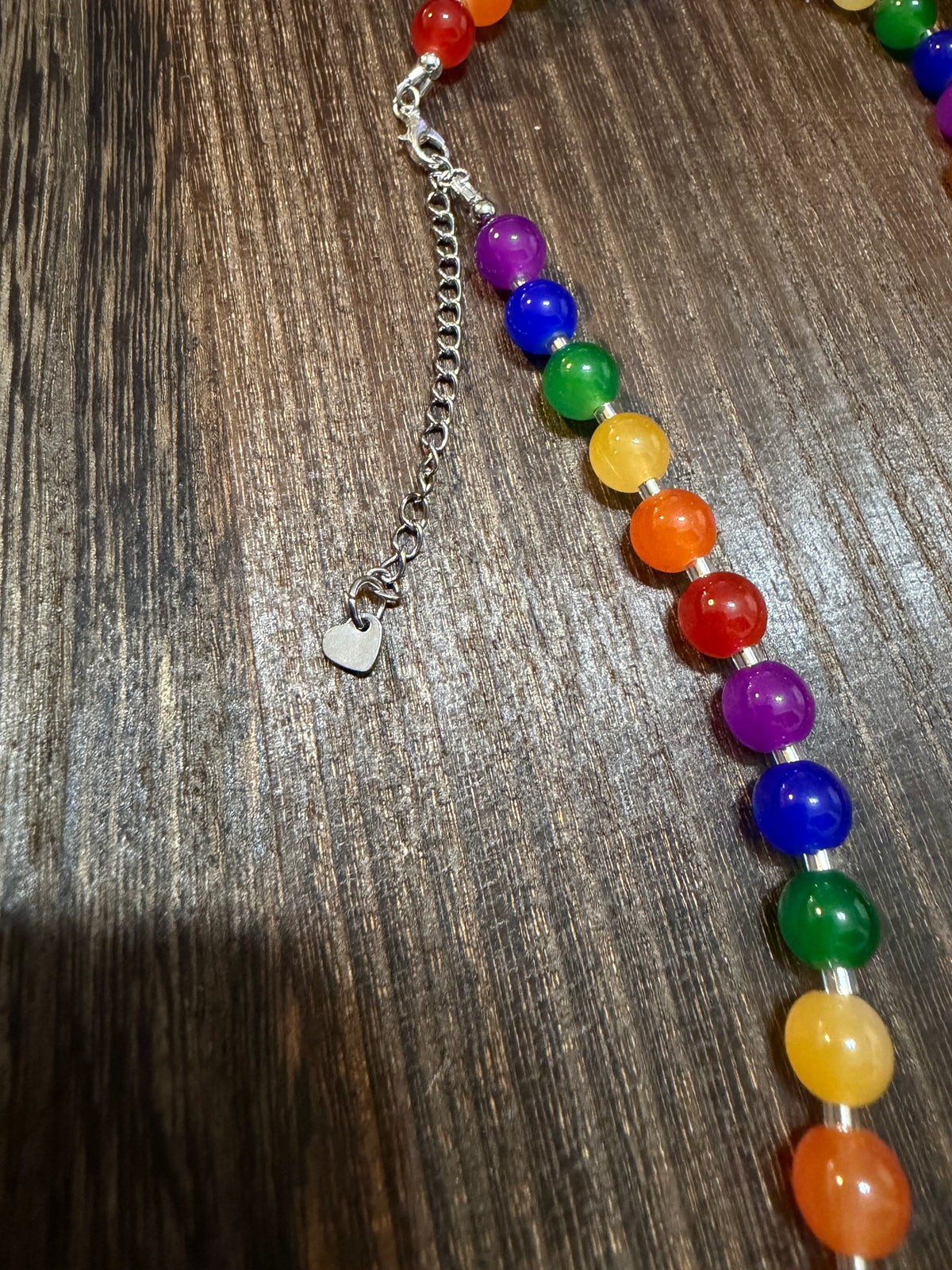 A close-up of a beaded necklace with translucent rainbow-colored round beads and a silver lobster clasp with a small heart charm, laid on a dark wood surface.
