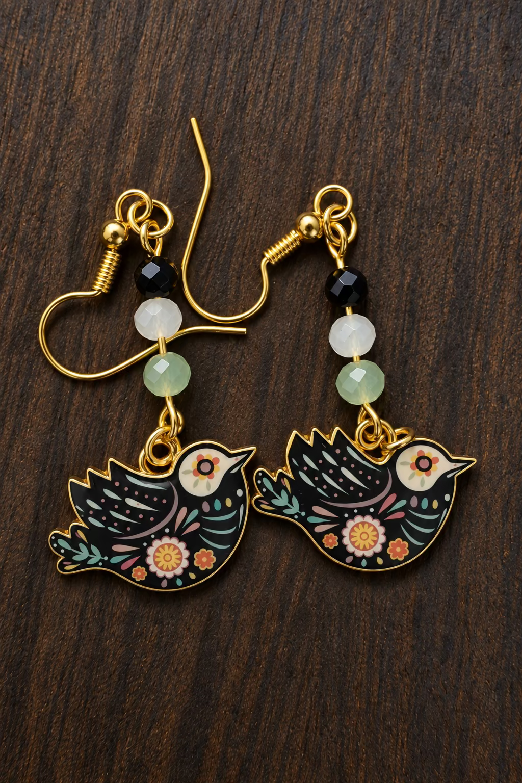 A pair of gold-tone dangle earrings with bird charms decorated in a colorful Day of the Dead style, resting on a dark wood background.