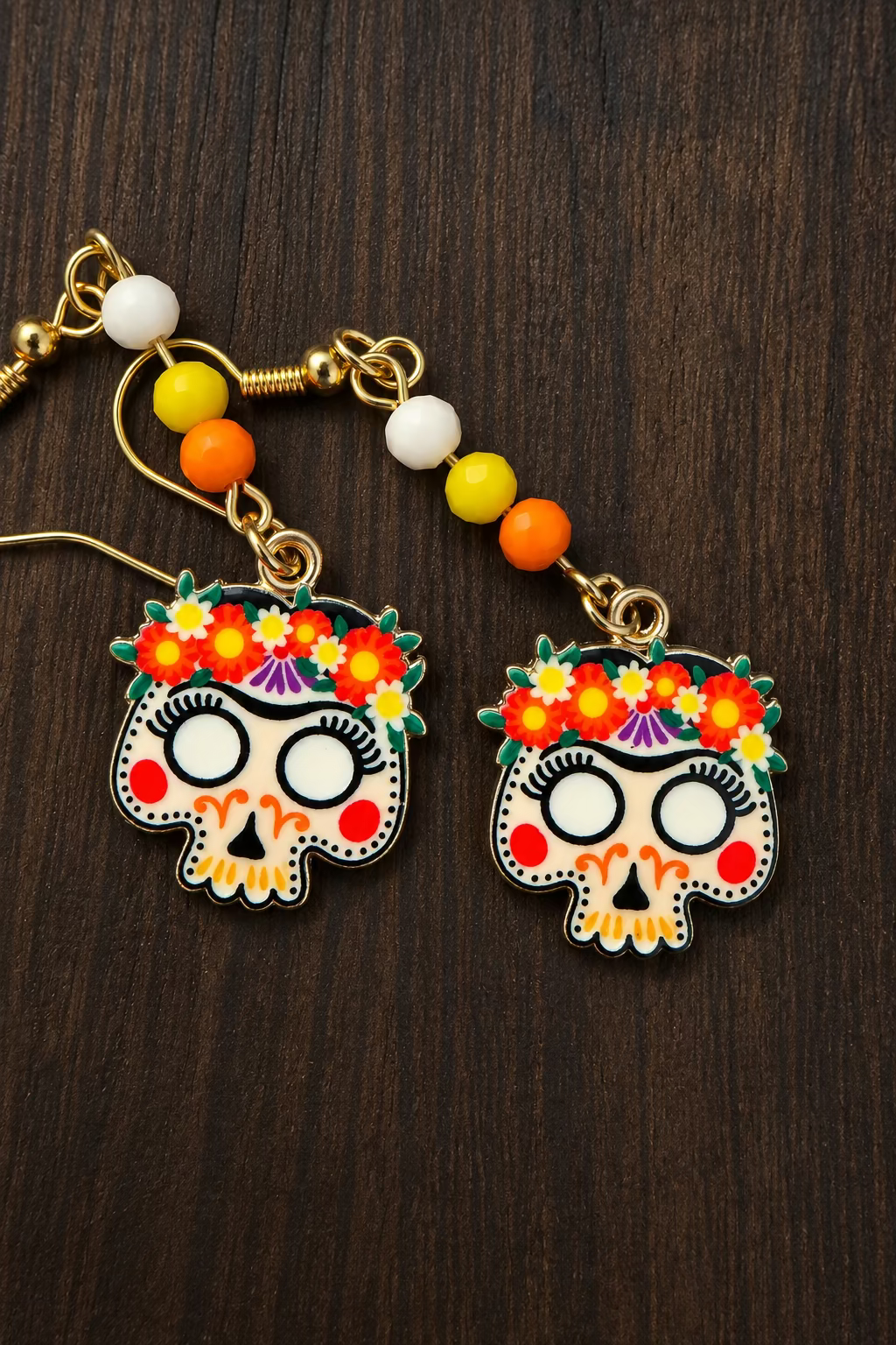 A pair of gold-tone dangle earrings with colorful sugar skull charms and floral crowns, resting on a dark wood surface.