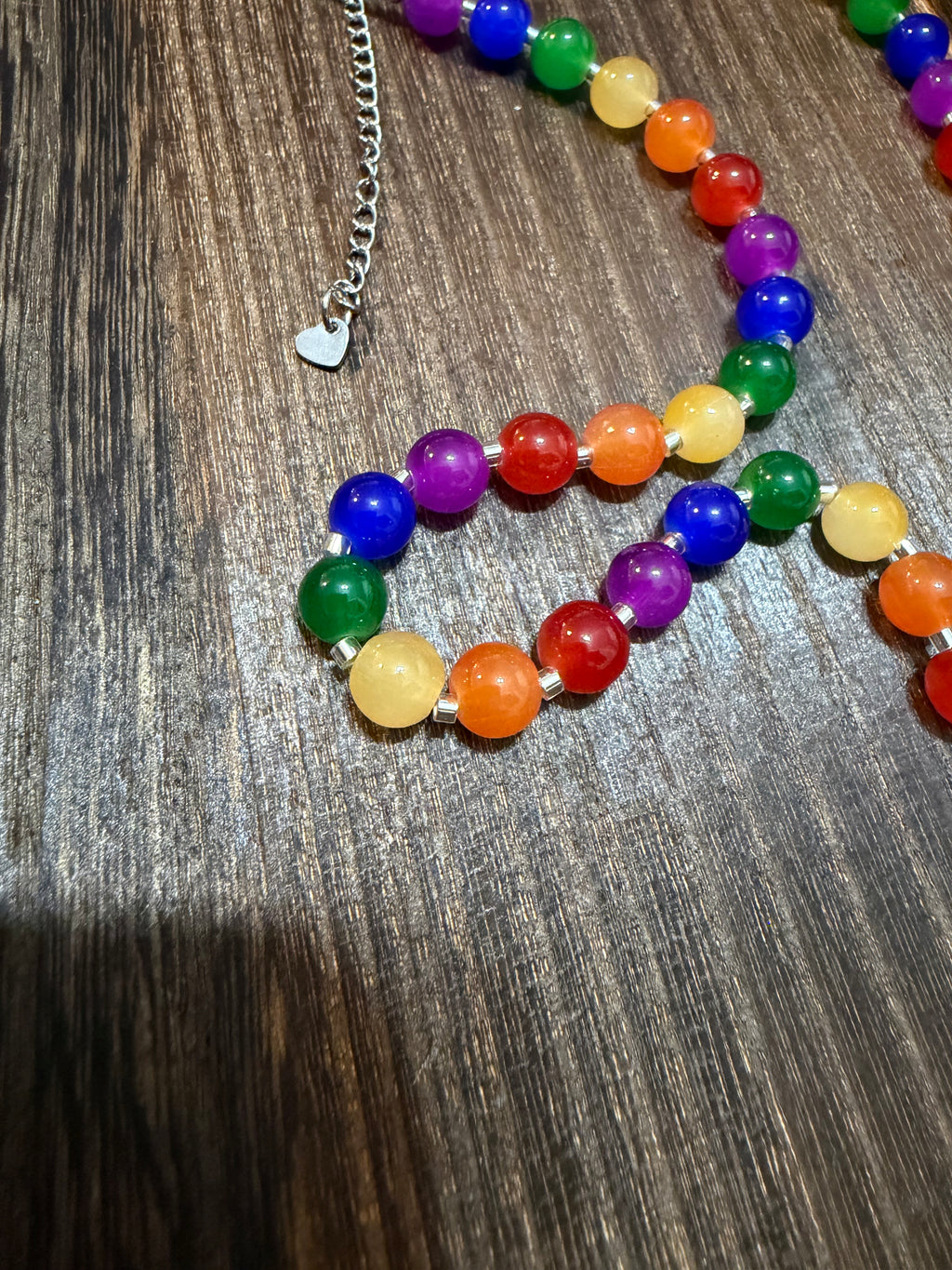 A rainbow beaded necklace made of translucent round gemstone beads in red, orange, yellow, green, blue, and purple, with a silver heart-shaped charm and chain, displayed on a dark wood surface.