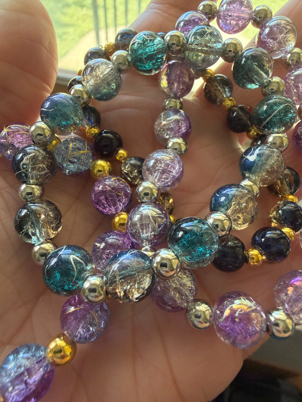 A close-up of a hand holding four beaded bracelets made of iridescent glass beads in purple, teal, blue, black, and clear, separated by small metallic gold and silver beads.