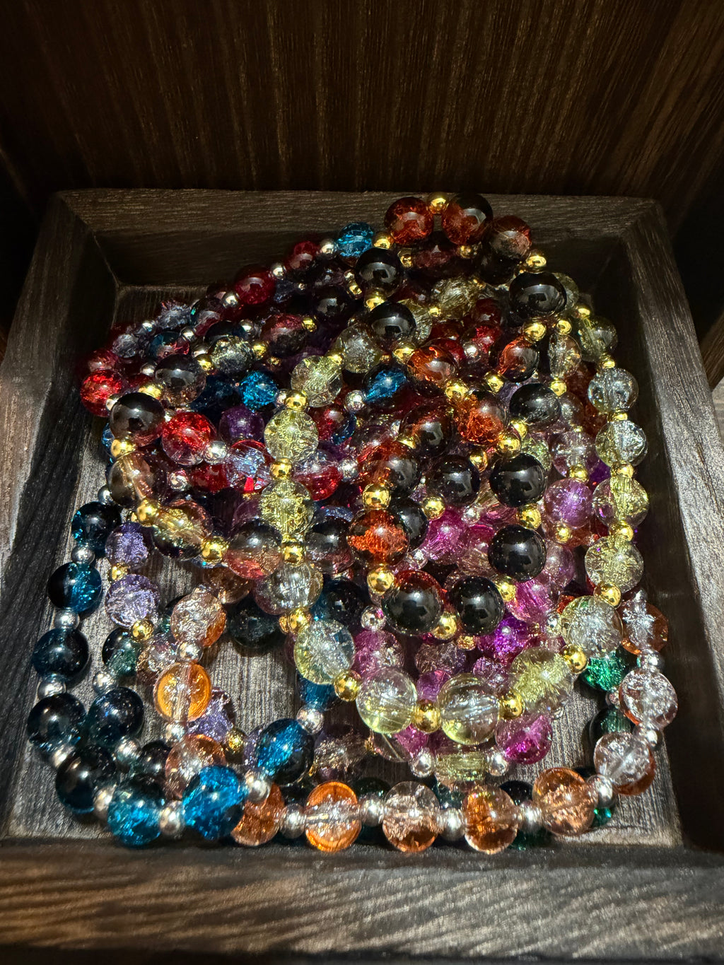 A pile of colorful beaded bracelets with crackle glass beads in many colors, arranged inside a dark wooden tray.