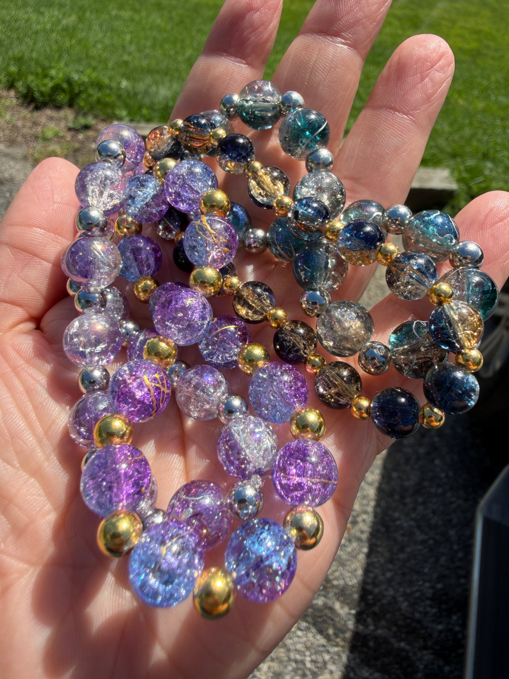 A close-up of a hand holding several crackle glass bead bracelets outdoors. The beads are primarily purple, blue, and clear with metallic gold accents, set against a background of green grass and pavement.  They are glittering.