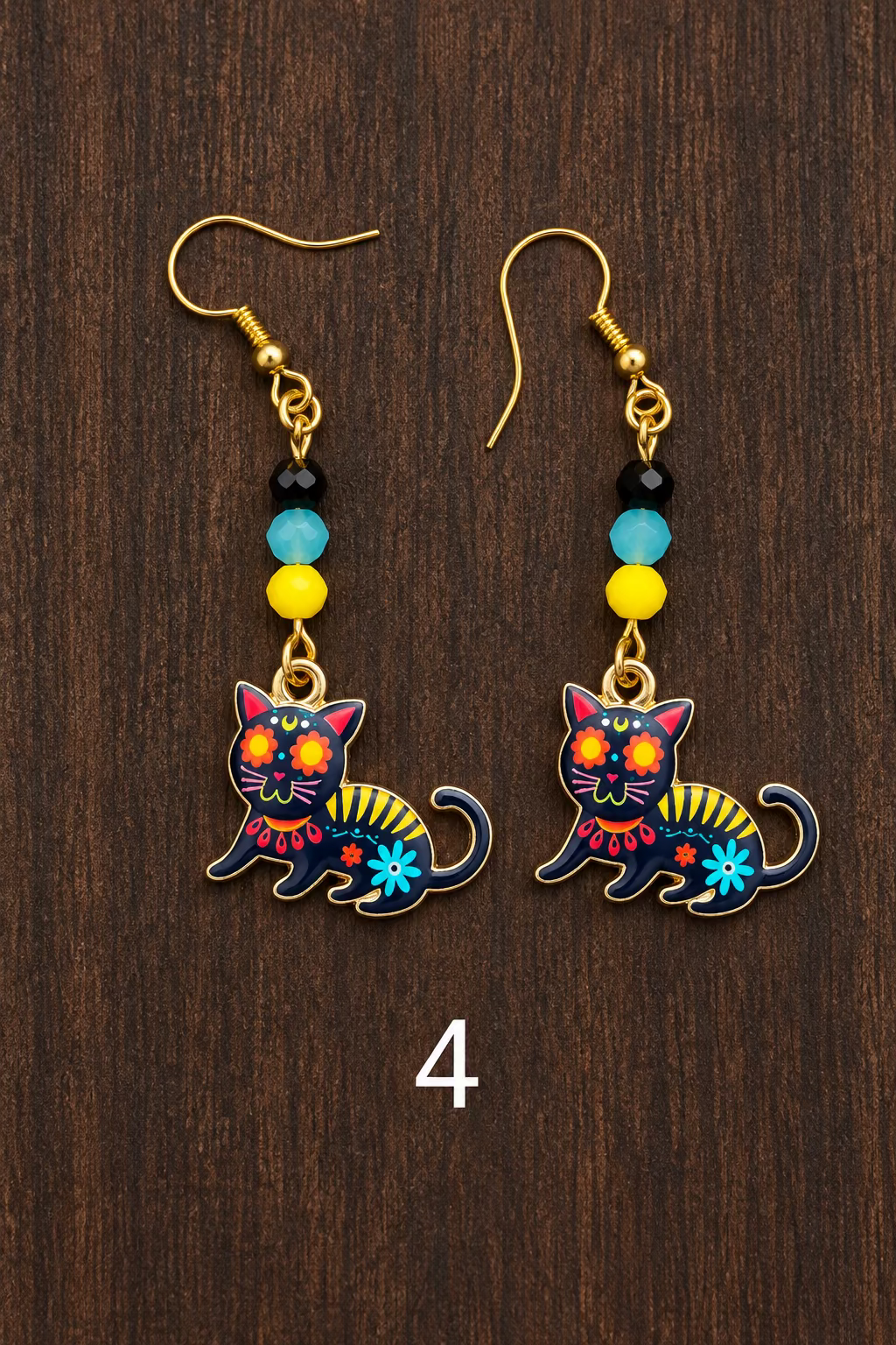 A pair of dangling earrings with gold-tone fishhook wires, small black and blue beads, and colorful cat charms decorated with floral and geometric patterns in red, yellow, blue, and white, displayed on a dark wood surface.