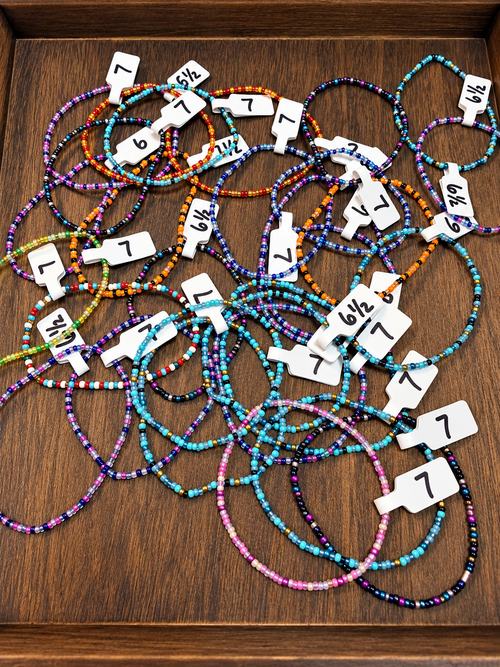 A pile of colorful beaded bracelets with white paper tags with their sizes on them, displayed on a dark brown wooden surface.