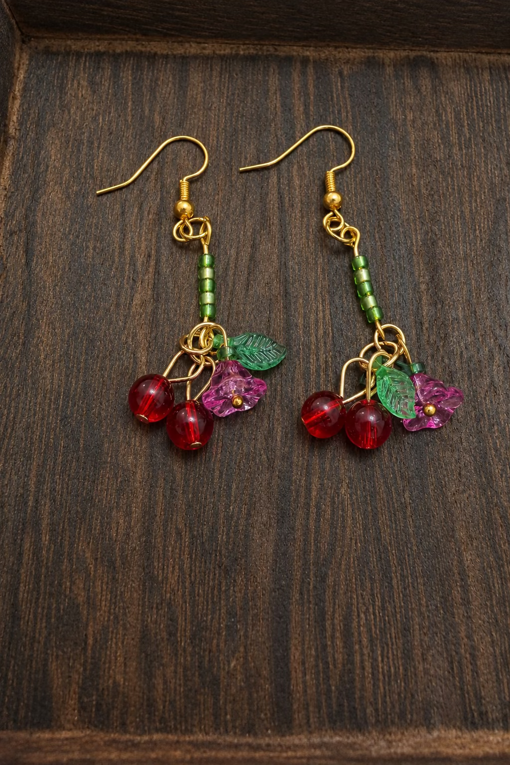 A pair of dangling earrings with gold-tone hooks, green beads, and clusters of red cherry beads with green leaf charms, displayed on a dark wooden surface.