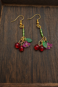 A pair of dangling earrings with gold-tone hooks, green beads, and clusters of red cherry beads with green leaf charms, displayed on a dark wooden surface.