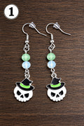 A pair of dangling earrings with skull charms wearing black witch hats, displayed on a dark wood surface. The earrings feature silver-toned metal hooks, light green and white beads, and white skull charms with black hat details.