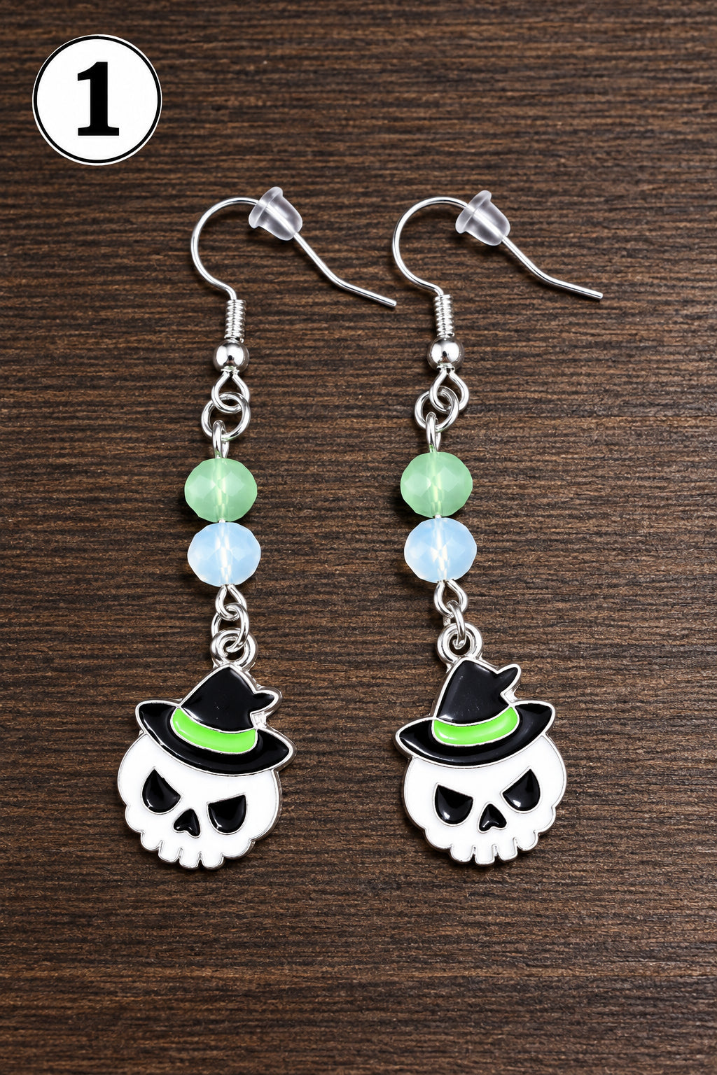 A pair of dangling earrings with skull charms wearing black witch hats, displayed on a dark wood surface. The earrings feature silver-toned metal hooks, light green and white beads, and white skull charms with black hat details.
