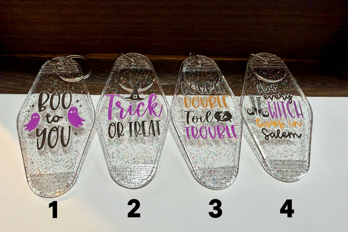 Four clear coffin-shaped acrylic keychains with glitter inside, each printed with a different Halloween-themed phrase: 'Boo to You,' 'Trick or Treat,' 'Double Toil & Trouble,' and 'Not Every Witch Lives in Salem.'.