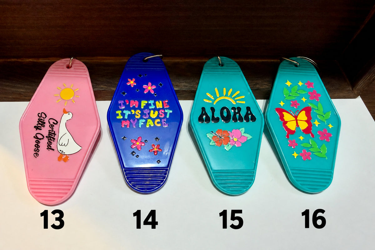 Four coffin-shaped plastic keychains lined up on a white surface. From left to right: pink with a white goose and yellow sun, blue with colorful flowers and the text 'I'M FINE IT'S JUST MY FACE', teal with 'ALOHA' and flowers, and turquoise with a yellow and pink butterfly and flowers.
