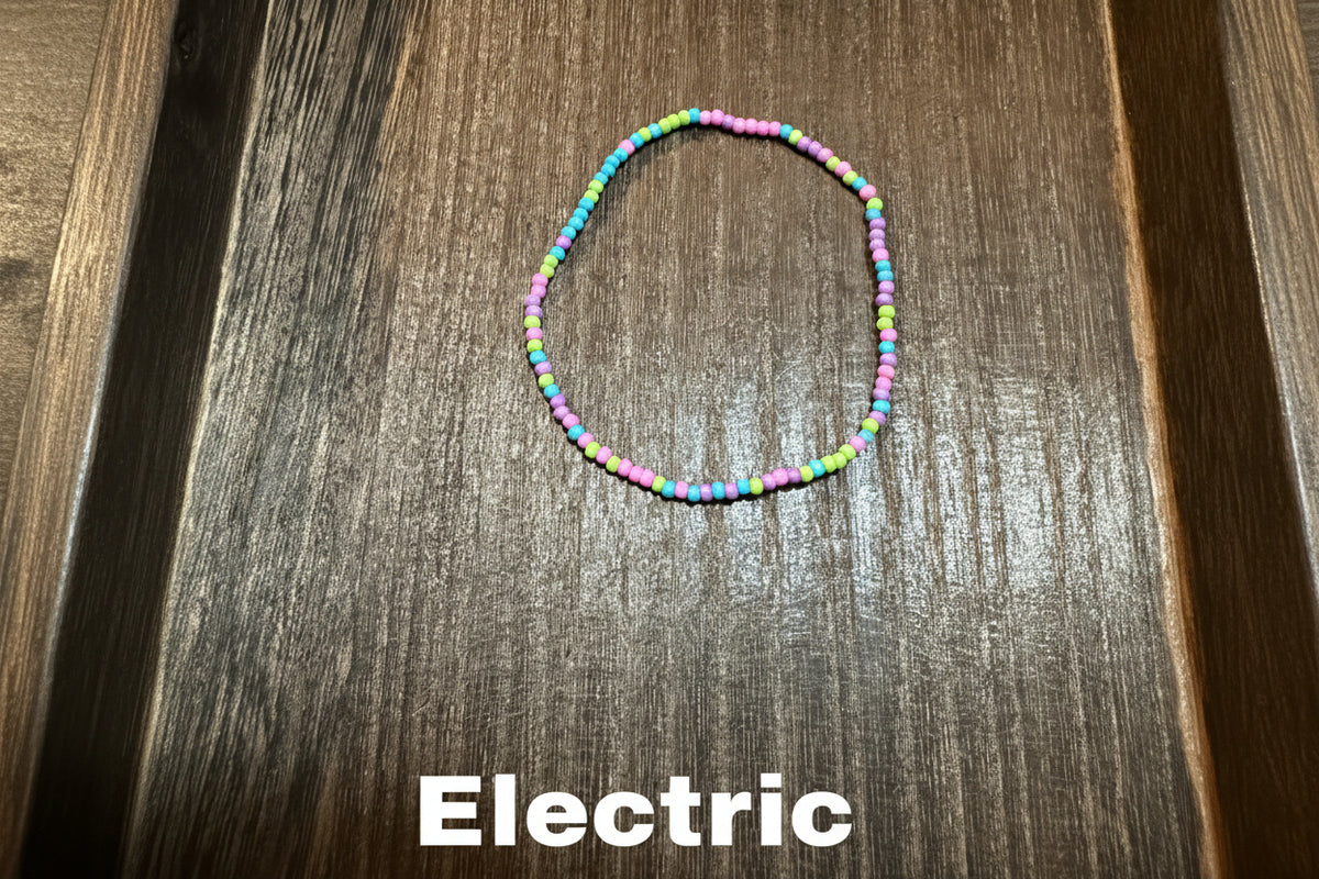 A small beaded bracelet made of tiny neon colored glass beads, arranged in a loose oval on a dark brown wood-grain surface.
