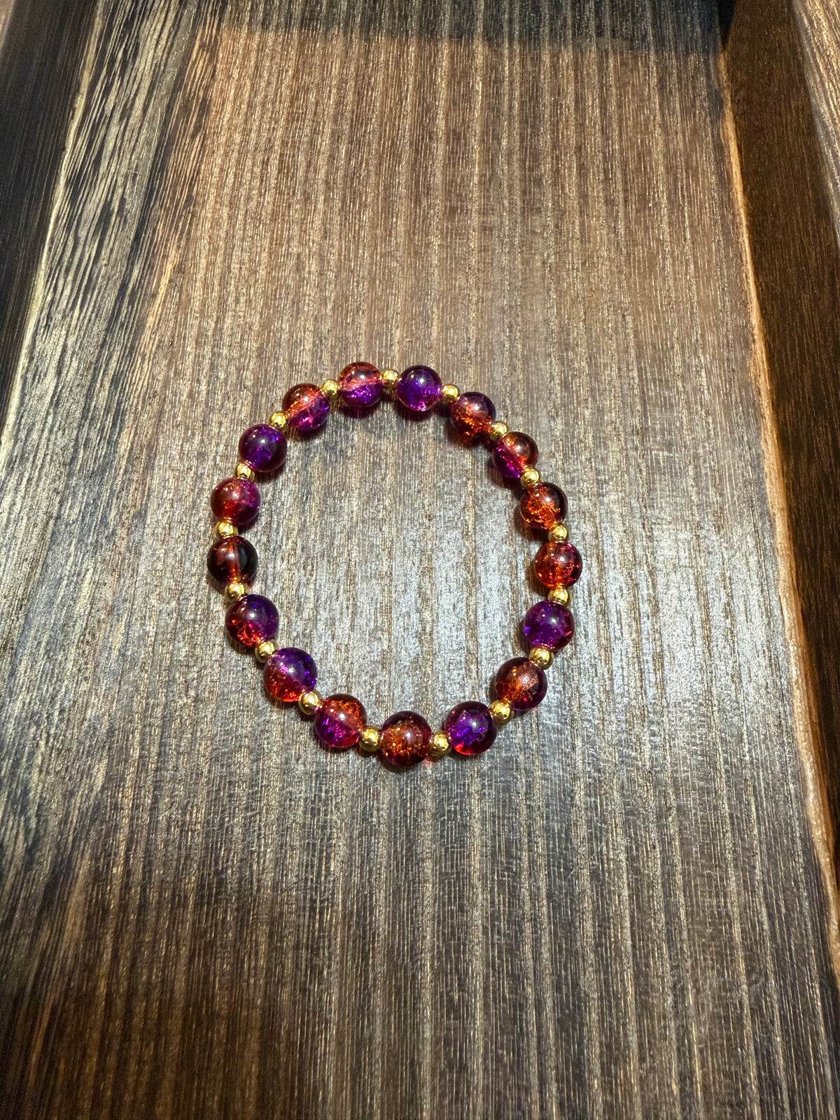 A beaded stretch bracelet with deep purple, reddish orange, and gold crackle glass beads separated by small gold spacer beads, displayed on a dark wood surface.