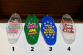 Four hotel keychains lined up on a white surface against a dark wood background. Each keychain has colorful printed text and glittery or clear plastic finishes.