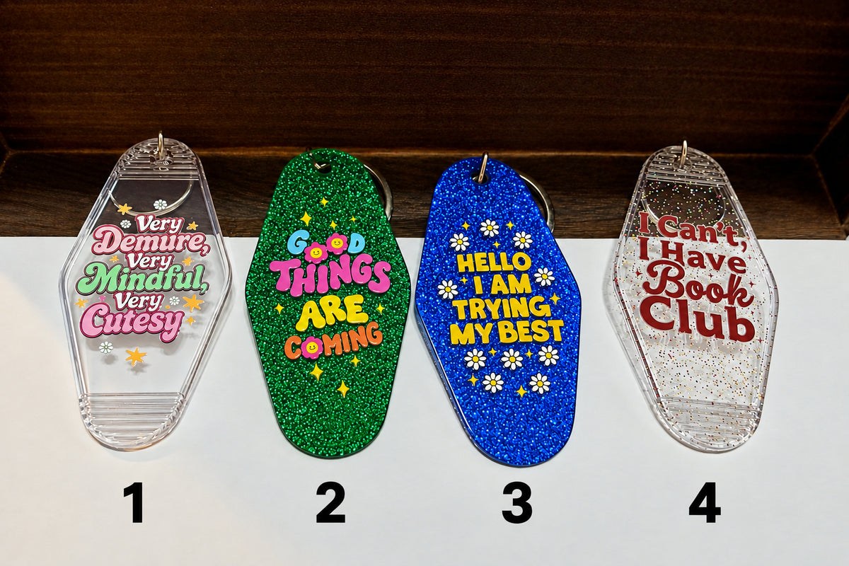 Four hotel keychains lined up on a white surface against a dark wood background. Each keychain has colorful printed text and glittery or clear plastic finishes.