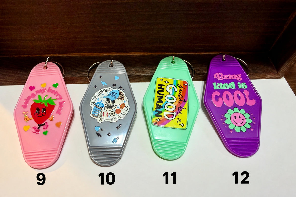 Four colorful coffin-shaped keychains lined up on a white surface against a dark wooden background. From left to right: a pink keychain with a strawberry graphic, a gray keychain with a skull and heart design, a mint green keychain with a yellow 'GOOD HUMAN' graphic, and a purple keychain with the text 'Being kind is COOL' and a smiley face flower.